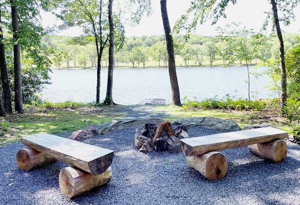 A serene lakeside setting with two wooden benches facing a fire pit surrounded by gravel.