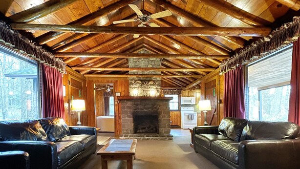 Interior de cabana de tronco com lareira de pedra, sofás de couro e ventilador de teto.