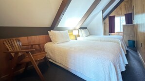 Ett sovrum med två sängar, en sänggavel i trä, en trästol och ett fönster med gardiner.
