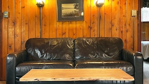 Um sofá de couro preto com uma mesa de centro de madeira em frente, apoiado em uma parede revestida de madeira, com duas luminárias fixadas na parede e um quadro emoldurado.