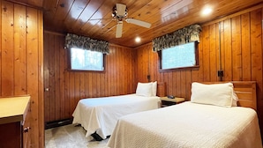 Quarto de cabana com duas camas, paredes de madeira, ventilador de teto e janela com cortinas.