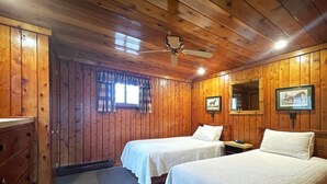 Una stanza con pareti e soffitto in legno, due letti con biancheria bianca, una ventola a soffitto, una finestra con tende e un comò.