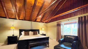 A bedroom with a wooden ceiling, a bed with a headboard, two bedside tables with lamps, a black leather chair, and a window with curtains.