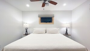 Una cama bien tendida con sábanas blancas en una habitación con paredes blancas, dos lámparas de noche y un ventilador de techo.