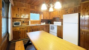 Une cuisine rustique aménagée dans une cabane en bois, avec une table en bois, un réfrigérateur, une cuisinière, un four à micro-ondes et une fenêtre avec des rideaux.
