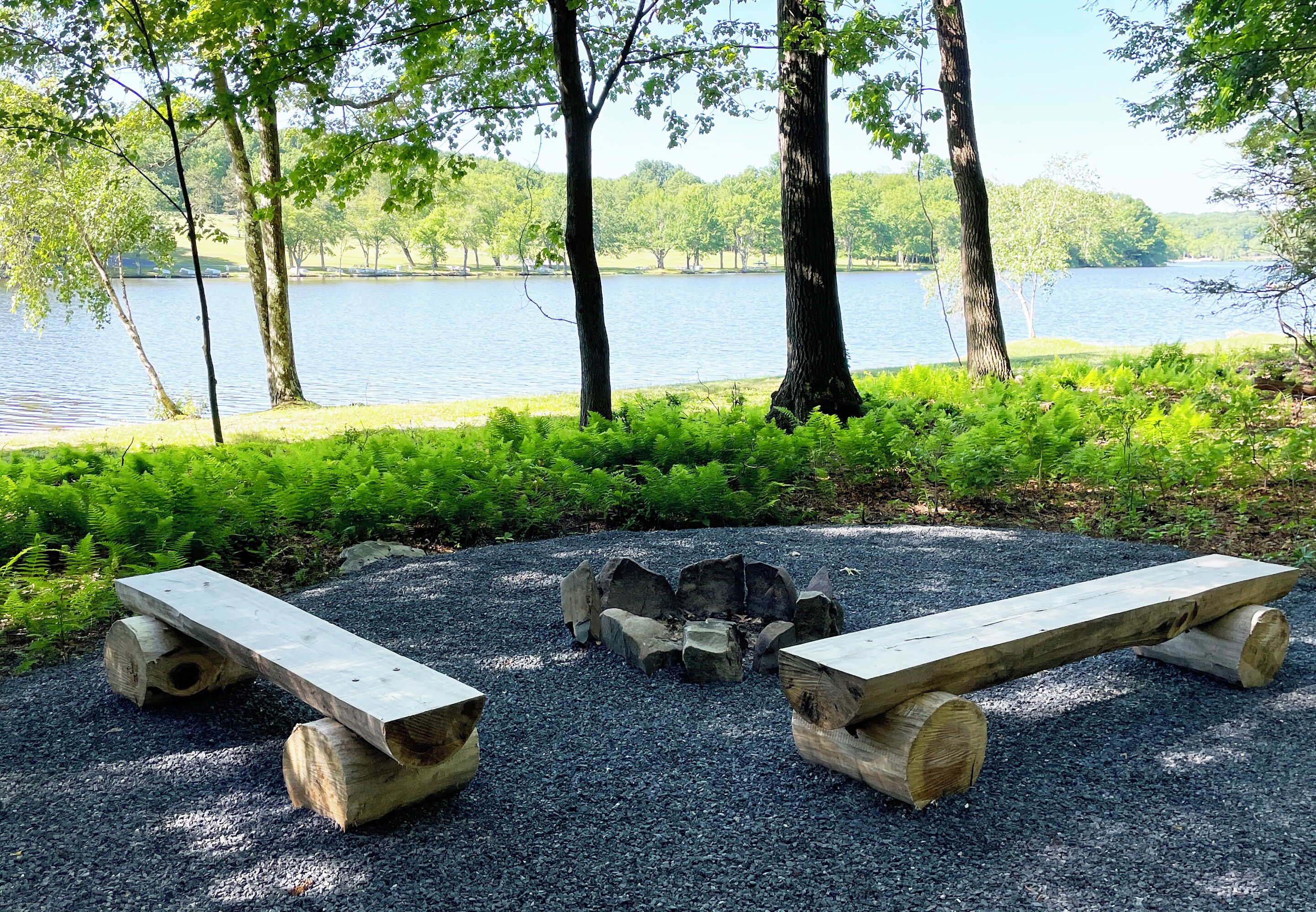 A wooden bench with a fire pit in the center, surrounded by gravel, with a lake and trees in the background.