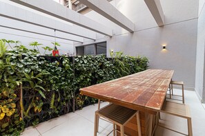 A modern outdoor dining area with a long wooden table and benches, surrounded by lush green plants and a white wall. - Vossa Bossa Think Republica (São Paulo)