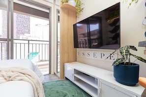 Una habitación de hotel moderna con un televisor grande montado en la pared, una cama con una colcha blanca, una alfombra verde, una planta pequeña en maceta y un balcón con vistas a la ciudad.