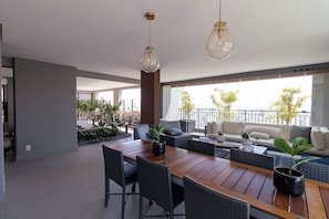A spacious living area with a wooden dining table, black chairs, and a sofa set. There are two hanging pendant lights, and the room has large windows with a view of the city. - Vossa Bossa Think Republica (São Paulo)