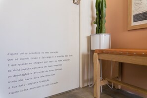 Una pared con un poema en portugués, un cactus en una maceta blanca y un banco de madera.
