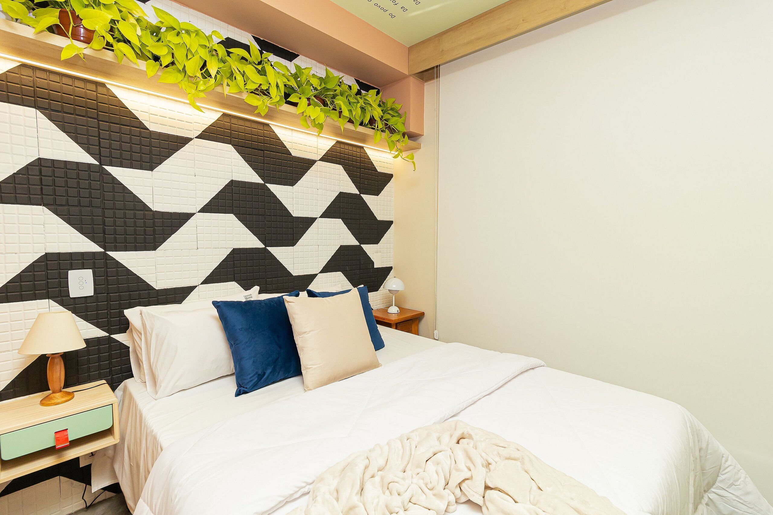 A modern bedroom with a geometric-patterned headboard, a bed with white bedding and blue pillows, a bedside table with a lamp, and a small nightstand.