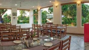 A restaurant with multiple tables and chairs, large windows with a view of greenery, and a red counter area. - Novel Resort by Urban (Ramnagar)