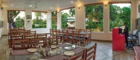 A restaurant with multiple tables and chairs, large windows with a view of greenery, and a red counter area.