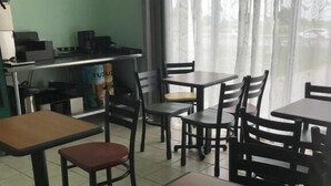 Ein Essbereich mit mehreren Tischen und Stühlen, eine Theke mit Registrierkasse und ein Fenster mit durchscheinenden Vorhängen.