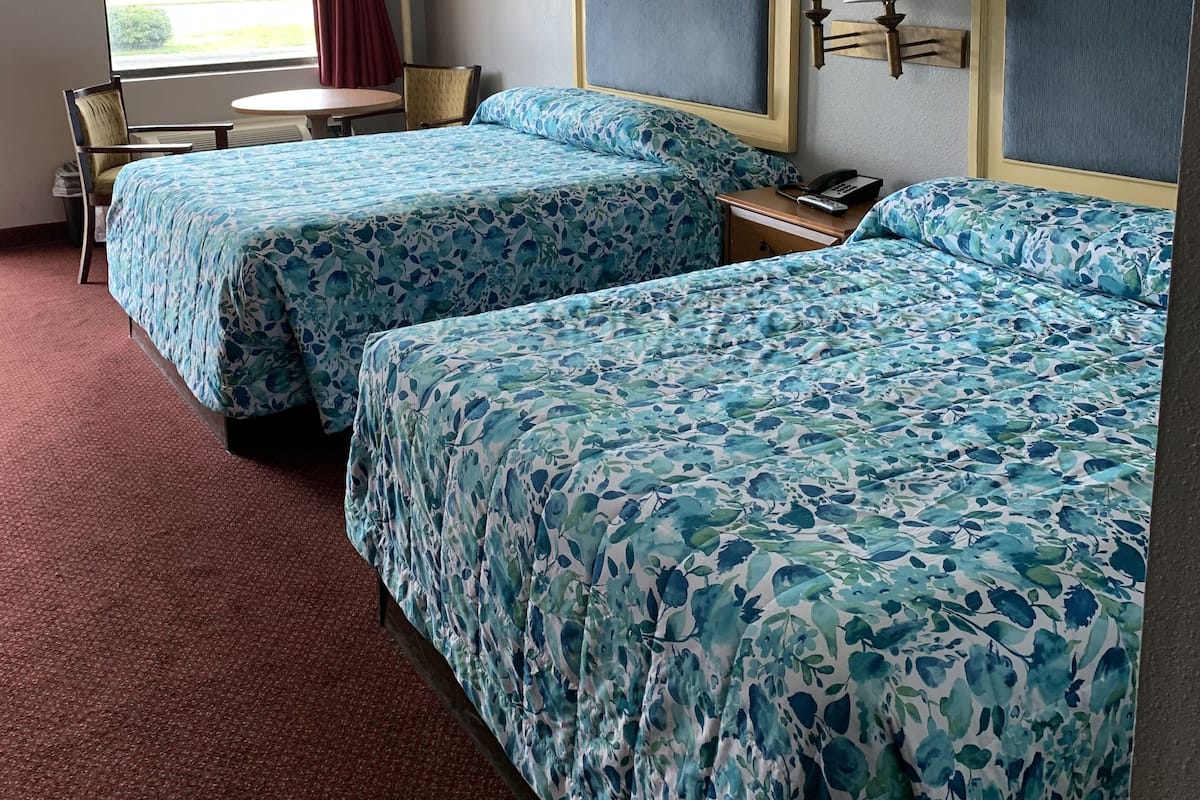 Quarto de hotel com duas camas, uma escrivaninha, uma cadeira e janela com vista para um edifício.