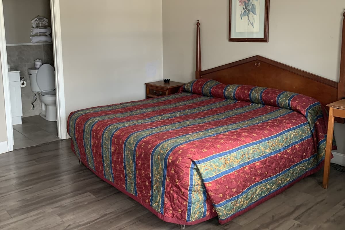 Quarto com cama de madeira, mesa de cabeceira, quadro na parede e banheiro visível através de uma porta aberta.