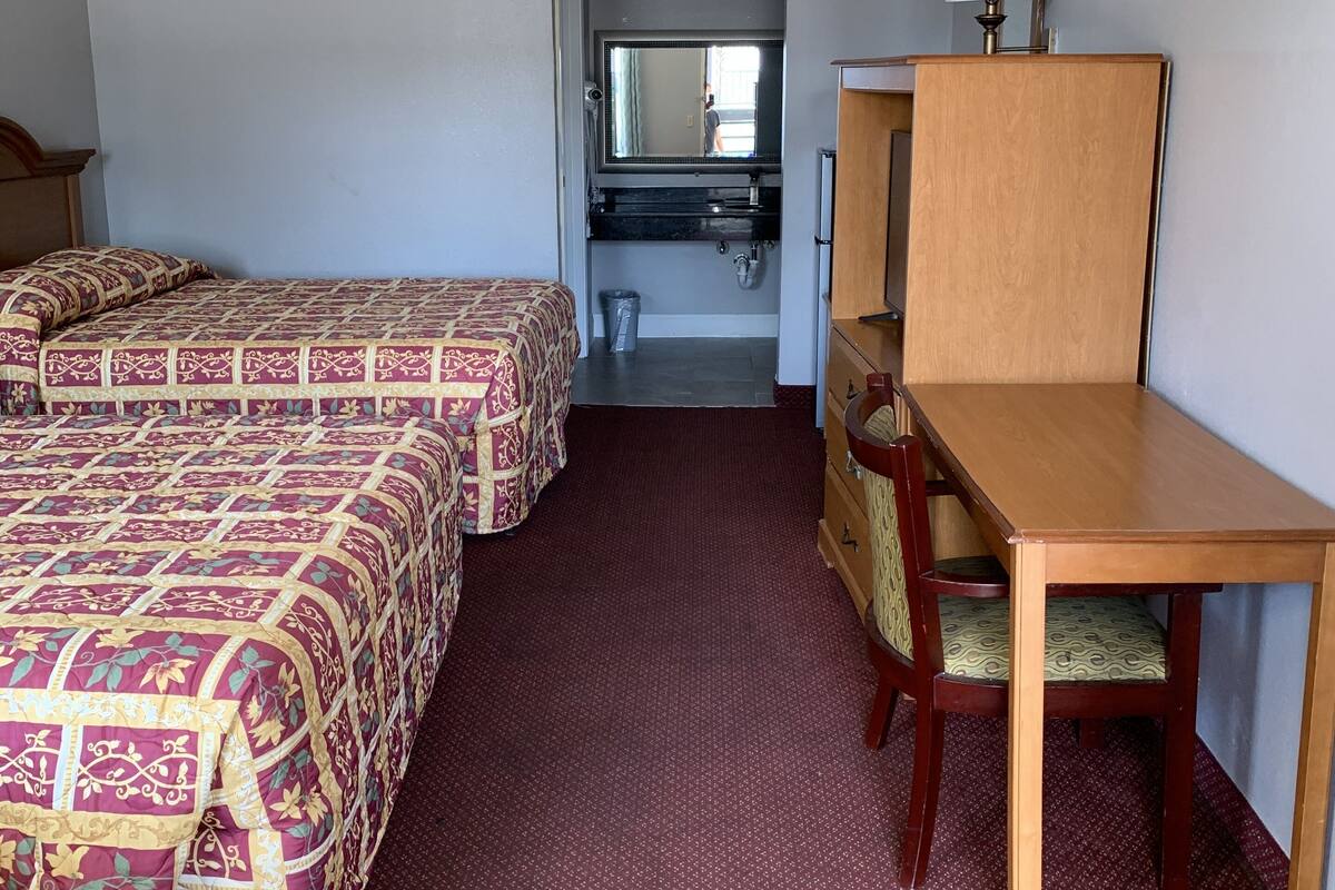 Quarto de hotel com duas camas, uma escrivaninha, uma cadeira, um espelho e um banheiro visível através de uma porta aberta.