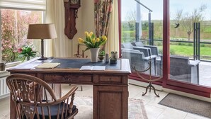 Una habitación con un escritorio de madera, una silla, un reloj y vistas a un jardín a través de un ventanal.