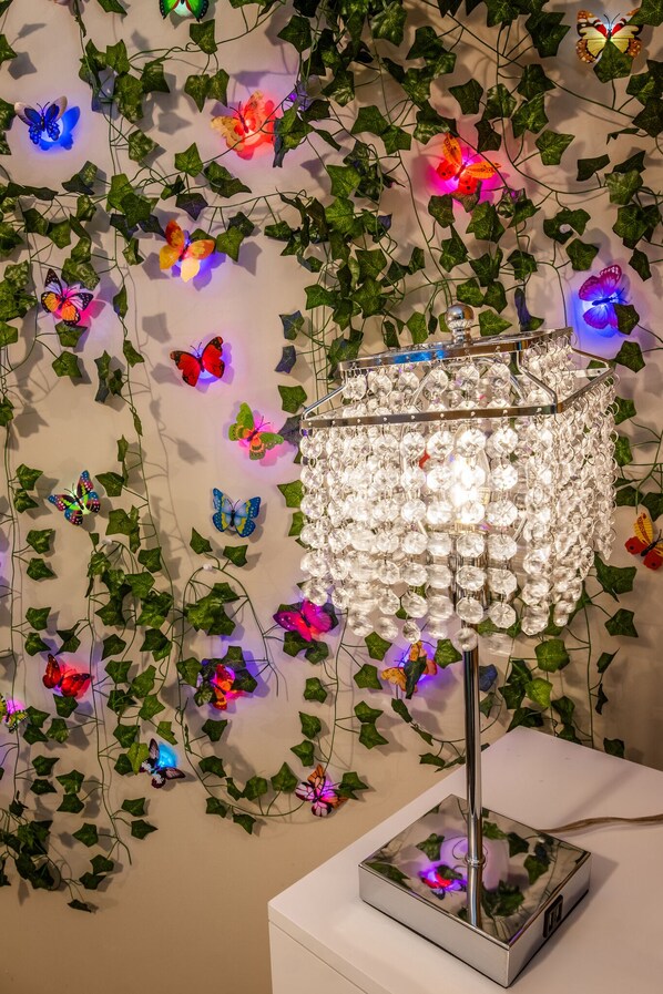 A chandelier with a square base and a metallic stand, surrounded by decorative butterflies and ivy on the wall.