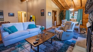 Una sala de estar acogedora con un sofá blanco, un sillón de cuero y una mesa de centro de madera. Se ve, al fondo, un comedor con una mesa y sillas.