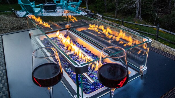 Una parrilla de cubierta con superficie de vidrio, con dos copas de vino tinto encima, rodeada por una terraza de madera y sillas.