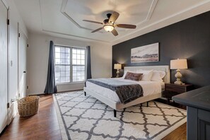Une chambre à coucher avec un lit, un ventilateur de plafond, une table de chevet avec une lampe, une fenêtre avec des rideaux et un tapis à motifs.