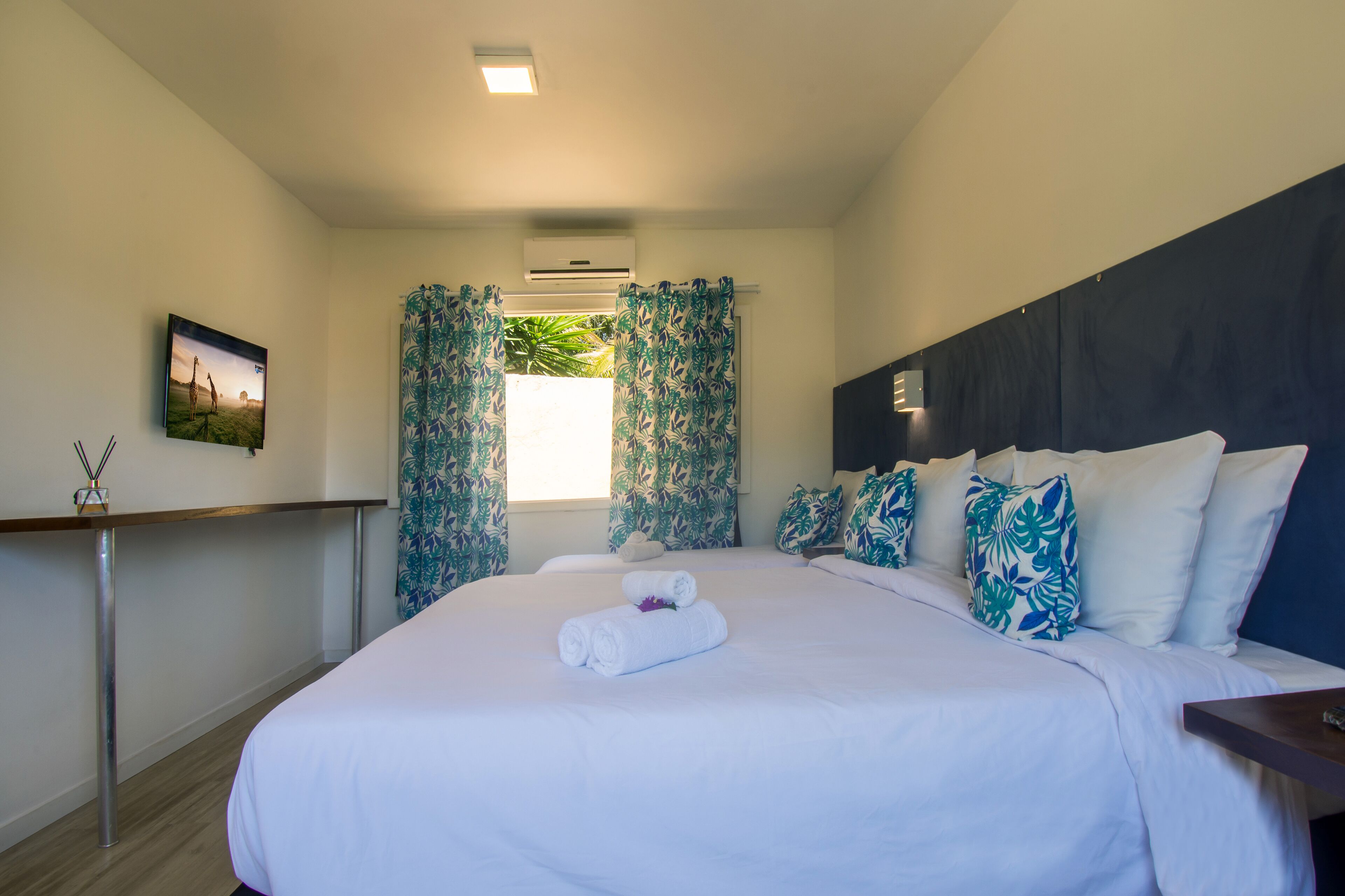 Una cama bien tendida con sábanas blancas y toallas enrolladas, una mesita de noche de madera, un aire acondicionado instalado en la pared y una ventana con cortinas estampadas.