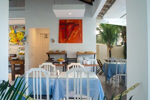 Uma sala de jantar com cadeiras e mesas brancas, um quadro grande na parede, e vista para o exterior.