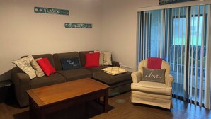 A living room with a sectional sofa, a small armchair, a wooden coffee table, and a ceiling fan. There are decorative signs on the wall and a view through a window with vertical blinds. - Winding Creek Condominiums (Clearwater)
