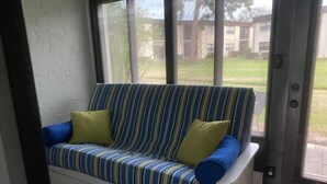 A corner sofa with striped upholstery and two pillows, positioned near a window with a view of a residential area. - Winding Creek Condominiums (Clearwater)