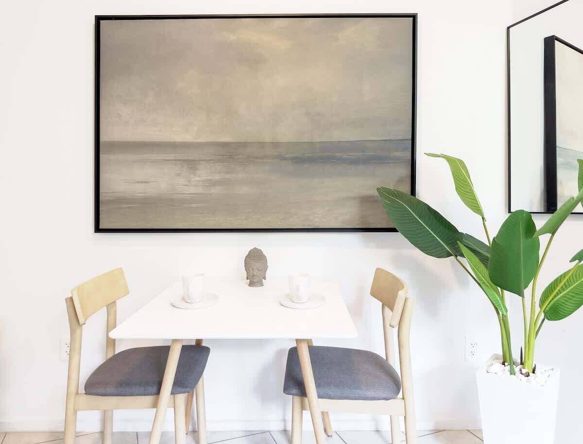 A dining area with a white table, two chairs, a Buddha statue, and a large abstract painting on the wall.