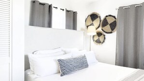 Een netjes opgemaakt bed met witte lakens en een gestreept kussen, een grijze hoofdbord en twee ronde decoratieve objecten aan de muur.