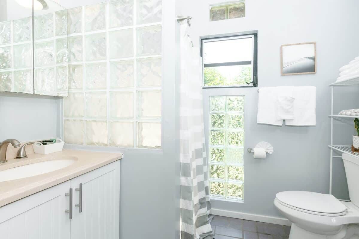 Un bagno con lavandino bianco, specchio e water. Si vede del verde attraverso una finestra.