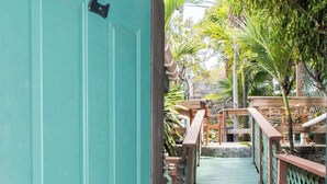 Een turquoise deur met een cijferslot, die uitkomt op een houten terras met een groene reling en een kleine brug.