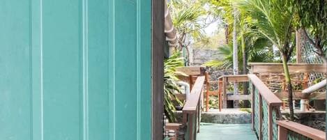 A turquoise door with a keypad lock, leading to a wooden deck with a green railing and a small bridge.
