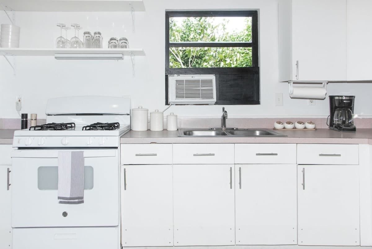 Una cocina con armarios blancos, estufa, pileta y ventana con vista a follaje.