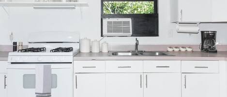 Una cocina con armarios blancos, estufa, pileta y ventana con vista a follaje.