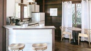 A compact kitchen with a central island, two stools, a dining table with chairs, and a window with curtains. - Oceanview Cottage | Walk to Beach & Restaurants (Cruz Bay)