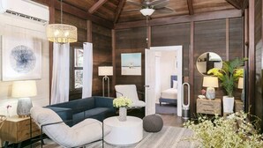 A modern living room with a dark blue sofa, white round coffee table, and a ceiling fan. There are framed pictures on the walls, a white chair, and a small side table with a lamp. - Oceanview Cottage | Walk to Beach & Restaurants (Cruz Bay)