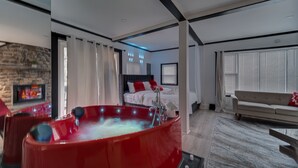 Ein modernes Hotelzimmer mit einer großen roten Whirlwanne, einem Steinkamin, einem Bett mit weißer Bettwäsche und einem Sofa.