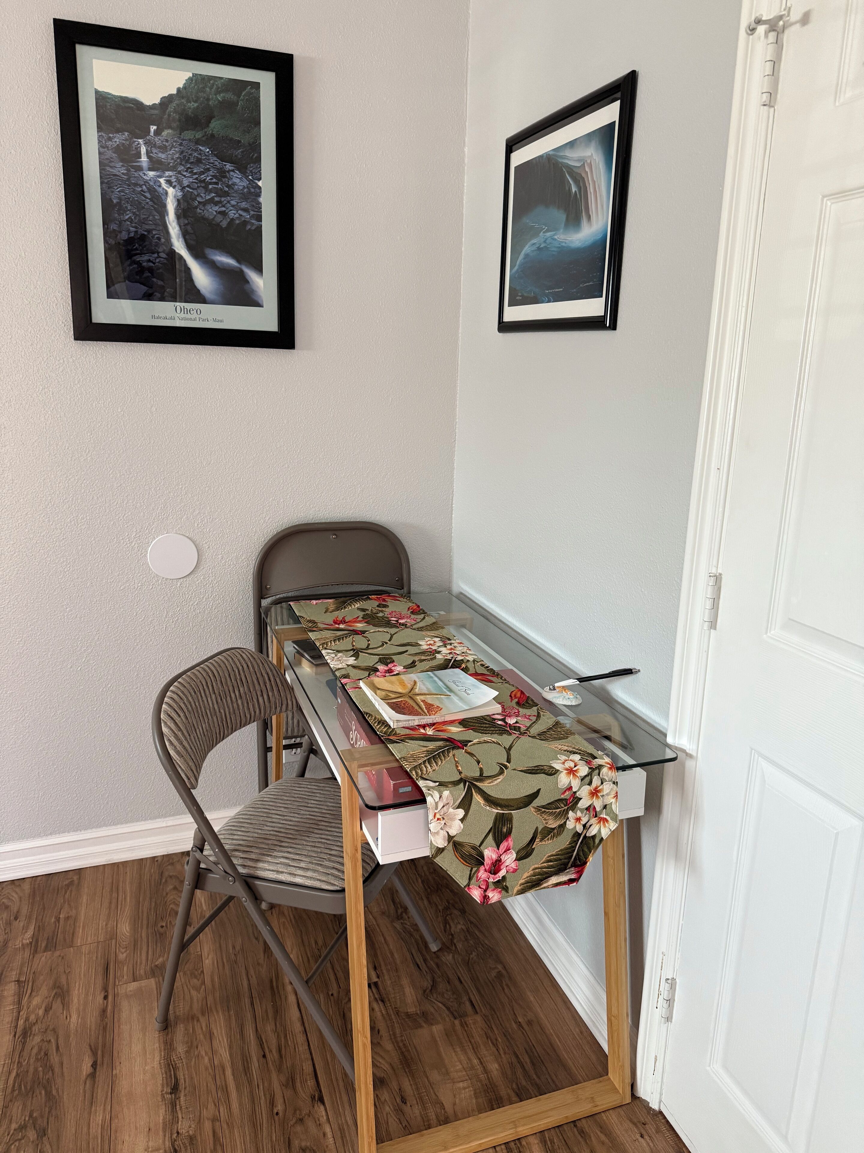 A small room with a wooden floor, a metal-framed desk with a floral tablecloth, a chair, and two framed pictures on the wall.