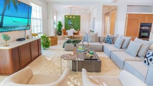 Un salón espacioso con una televisión de pantalla plana grande, un conjunto de sofás, una mesa de centro y un comedor con una pared verde y una persona sentada a la mesa.