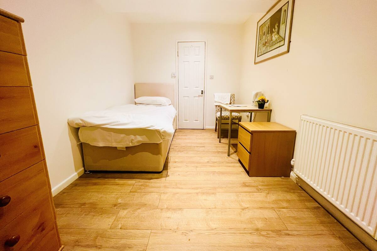 A small, well-lit room with a bed, a desk, a chair, a dresser, and a radiator.