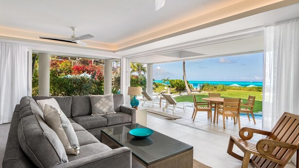 Una sala de estar moderna con un sofá gris, una mesa de centro de vidrio y una silla de madera. Se ve una ventana grande que ofrece vistas al océano y un área de patio con mobiliario exterior.