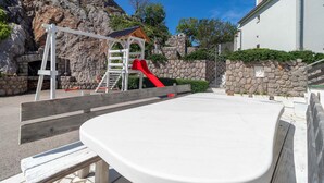 Un parque infantil con un tobogán y una estructura de escalada, frente a un muro de piedra y un edificio de fachada blanca.