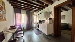 Een compacte keuken met witte kastjes, een magnetron en een koffiezetapparaat. Er is een eethoek met een tafel en stoelen, en een deur naar een woonruimte.