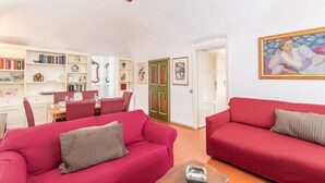 Ein Wohnzimmer mit roten Sofas, eine Essecke mit Tisch und Stühlen sowie ein Bücherregal voller Bücher und dekorativer Gegenstände.