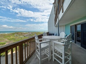 A balcony with a view of the beach and ocean, white chairs, and a table.