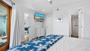 Un dormitorio con una cama, un televisor montado en la pared, un tocador y una puerta que da a otra habitación.