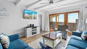 Amplia sala de estar con ventilador de techo, televisor grande y vista al mar a través de un ventanal corredero.